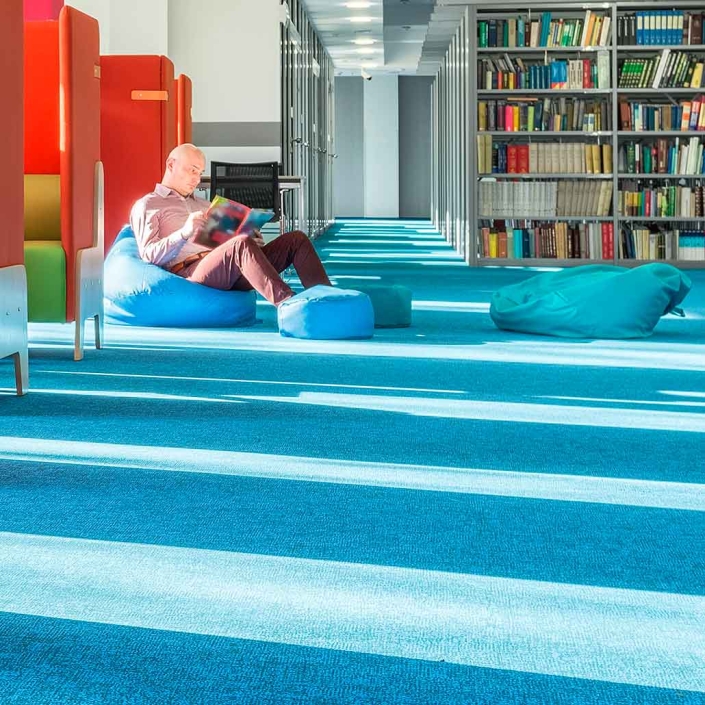 Alfombra para tráfico intenso en color azul colocada en zona de lectura de una biblioteca con un hombre leyendo sobre un taburete