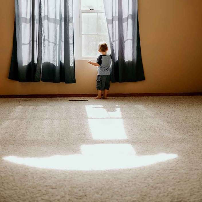 Niño descalzo sobre una alfombra residencial en color claro asomado en una ventana