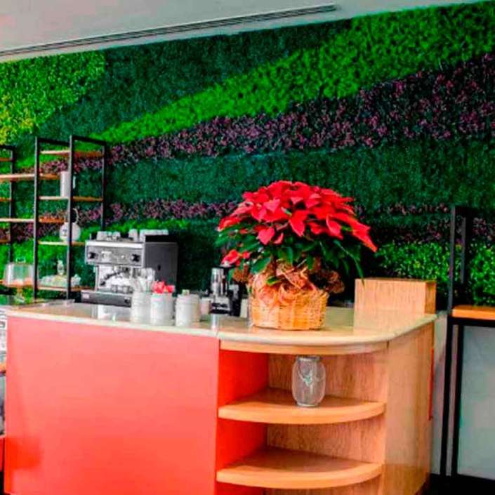Muro interior recubierto con muro verde artificial en tonalidades verdes y moradas colocado en cafetería con mobiliario de madera y anaqueles metálicos